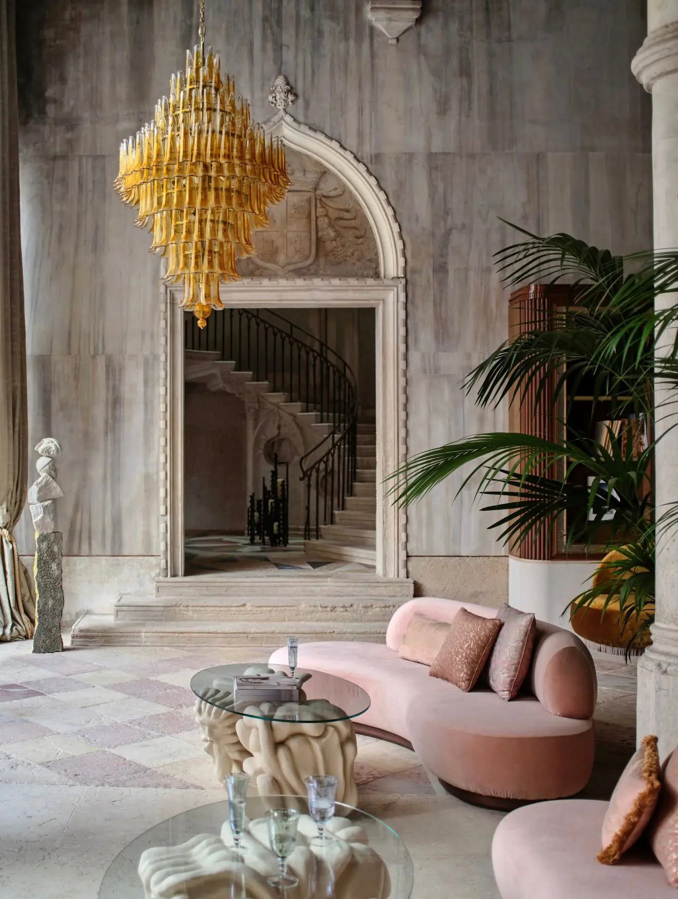 A marble palazzo lounge with an amber chandelier, blush curved sofa and potted plants